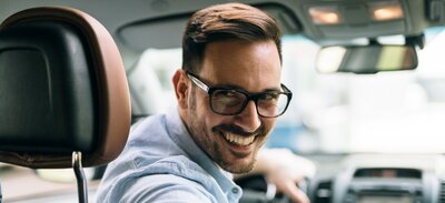Junger lächelnder Mann mit Brille sitzt in seinem Dienstwagen und fährt rückwärts.